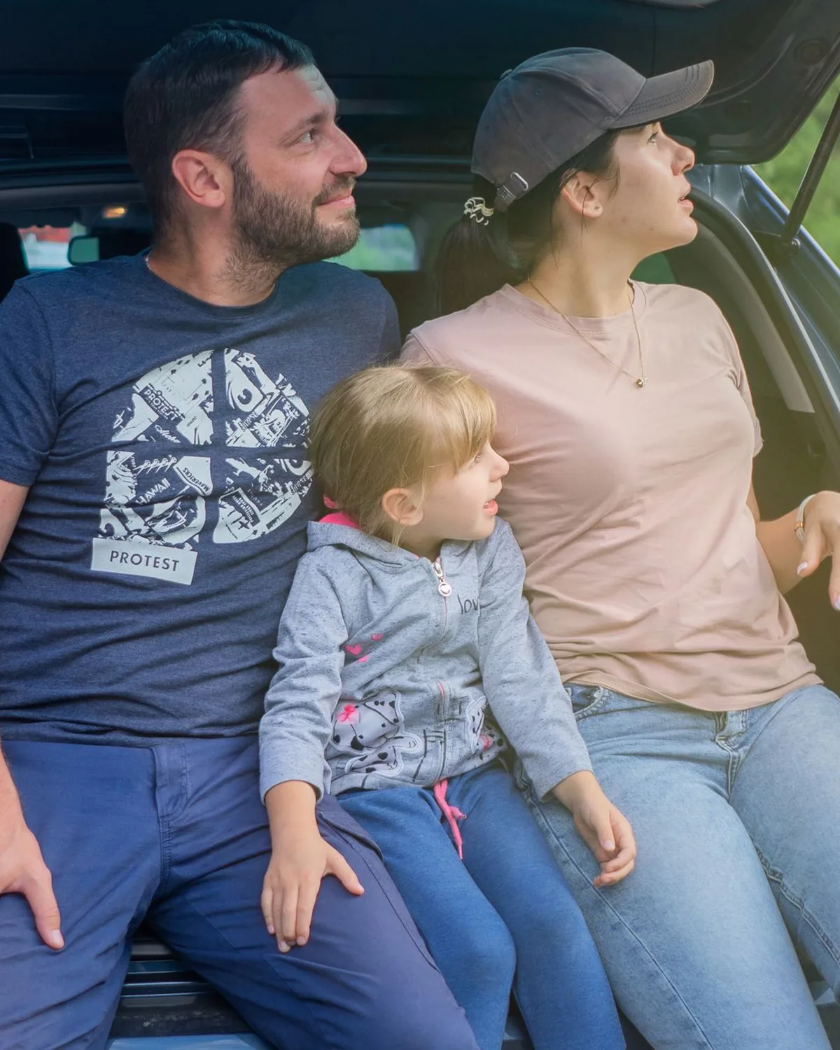 A family with a small child sitting together in the open boot of their car on a trip