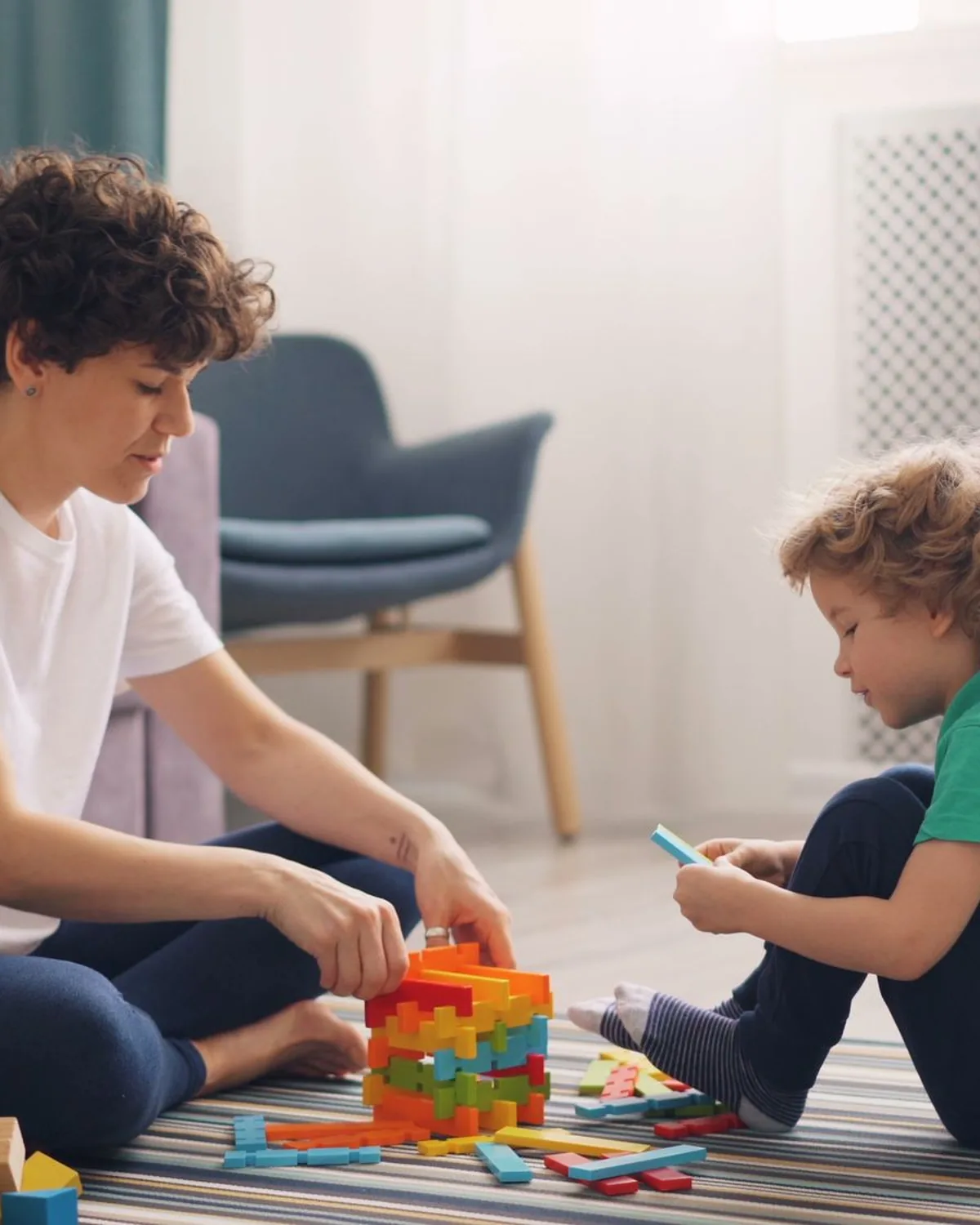 A caregiver and a young boy building with coloured blocks on the floor of a bright living room
